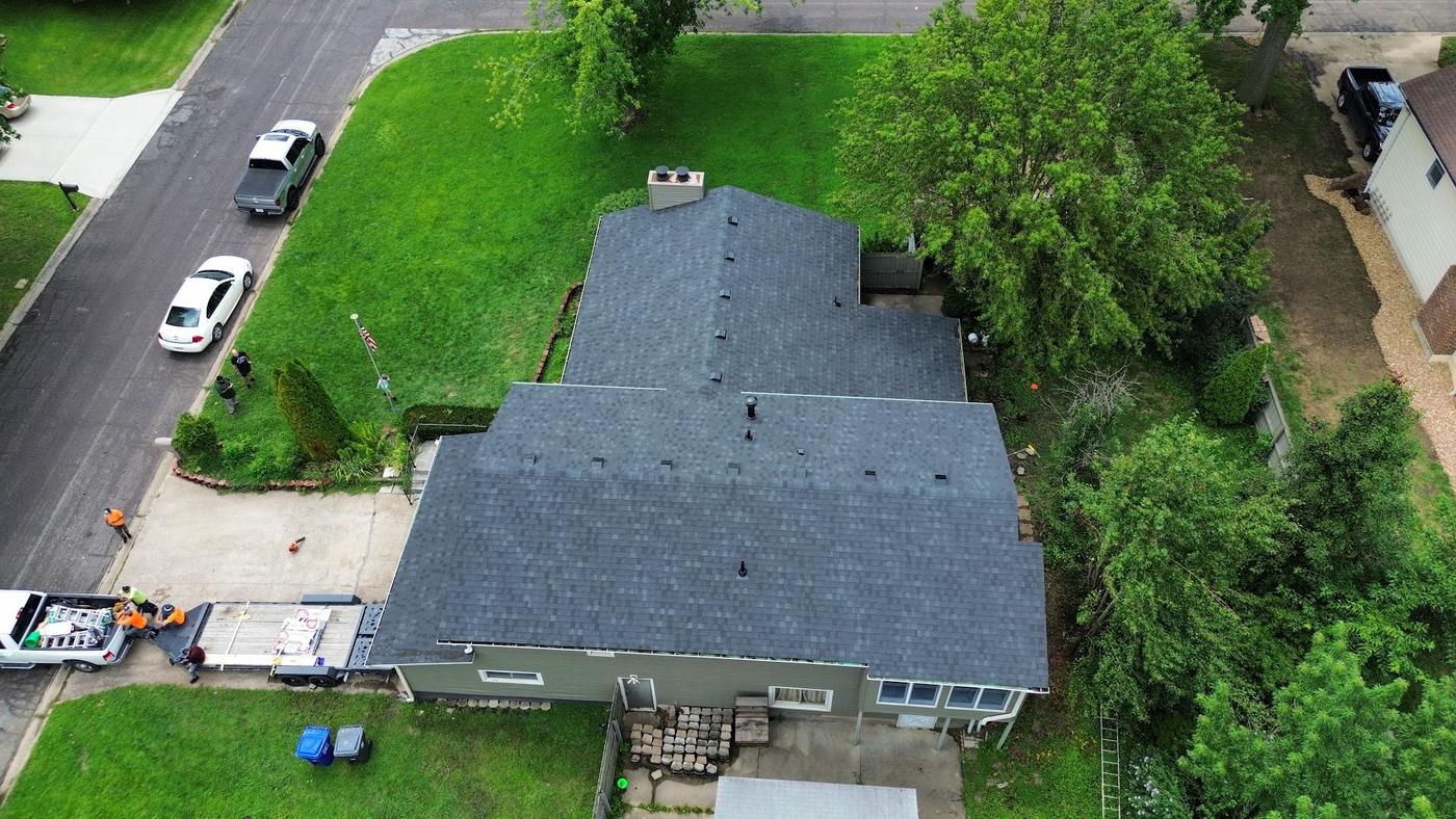 A drone view of a crew actively working on a shingle roof, materials staged on a trailer, workers in orange safety gear